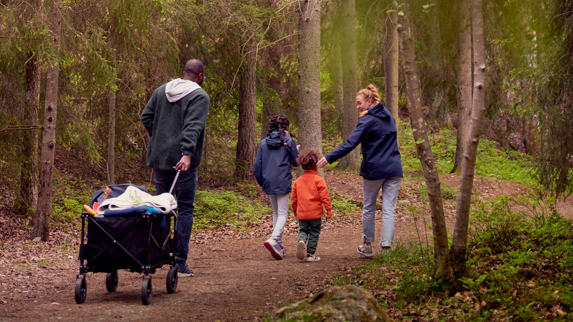 Familj med två barn promenerar i skogen och drar en campingvagn efter sig. Sverige.