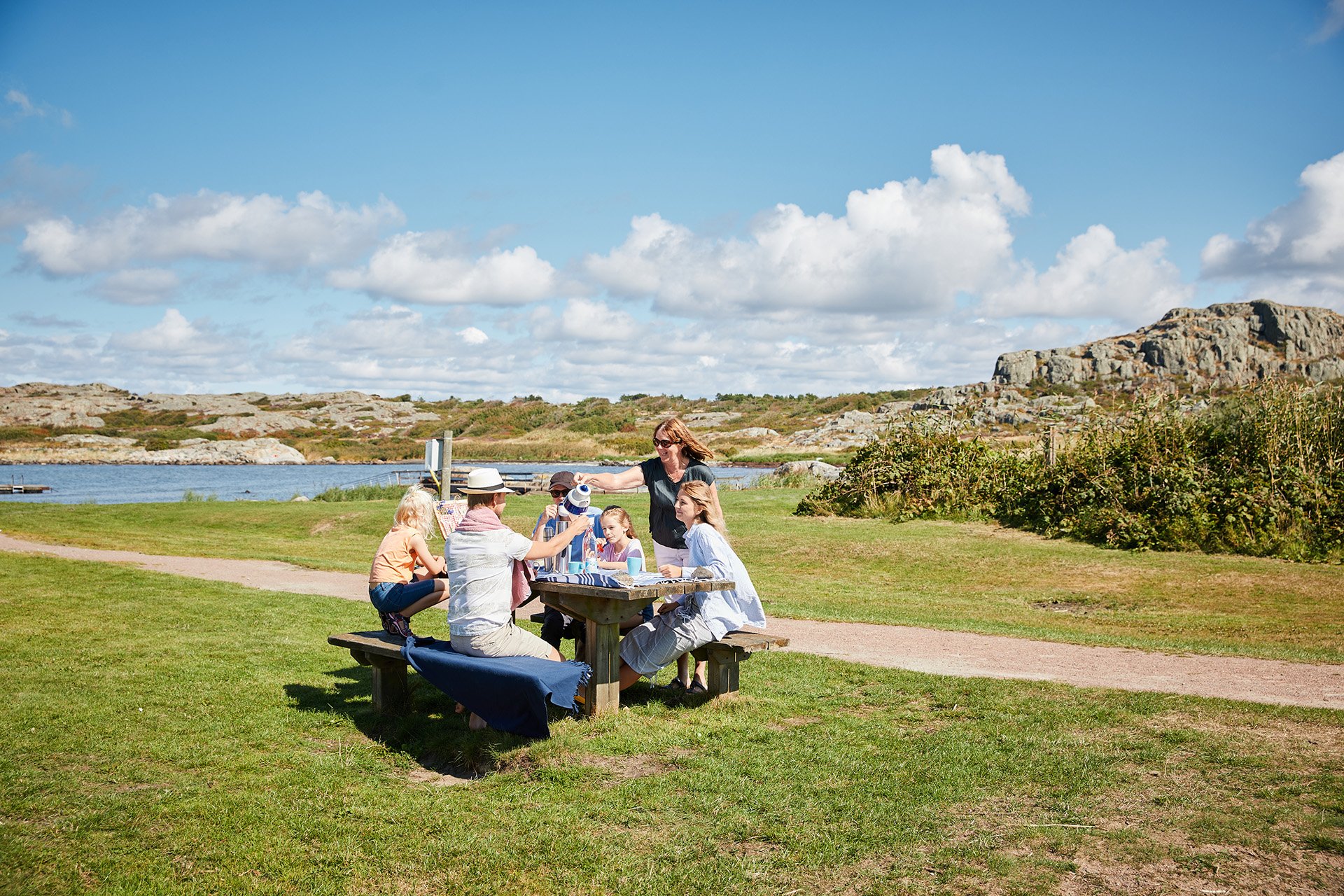 Två vuxna och fyra barn som sitter runt ett picknickbord i skärgårdsmiljö